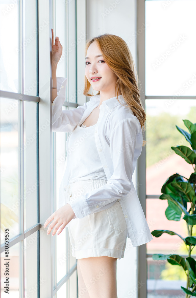 Asian beautiful female girls smile standing by the window, Cheerful young woman happy in the living room, feeling so relaxed at home weekend lifestyle in the morning. Healthcare concept Asian beautiful female girls smile standing by the window, Cheerful young woman happy in the living room, feeling so relaxed at home weekend lifestyle in the morning. Healthcare concept