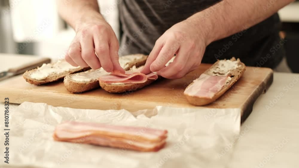 Caucasian male carefully placing bacon strips on sliced baguette in the kitchen. Caucasian male carefully placing bacon strips on sliced baguette in the kitchen.