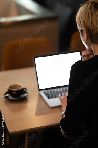 A professional woman is seen from behind working intently on her laptop in a stylish, modern cafe. A coffee cup is visible beside her, enhancing the work ambiance.