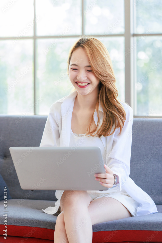 Happy young asian beautiful woman smiling sitting relaxed on cozy sofa in living room using laptop computer, shopping online, reading news, browsing internet at home, Girl using digital technology Happy young asian beautiful woman smiling sitting relaxed on cozy sofa in living room using laptop computer, shopping online, reading news, browsing internet at home, Girl using digital technology