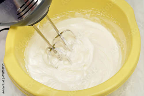 Mixing egg white cream with electrical hand mixer in the yellow plastic bowl. Close-up. Selective focus.