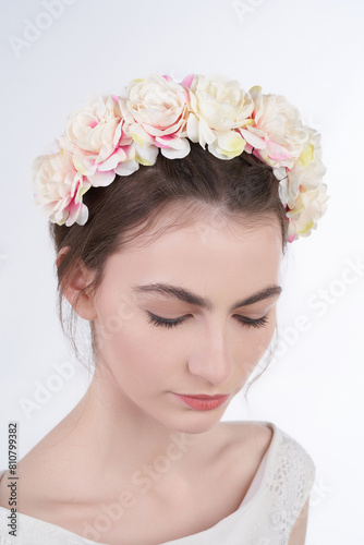 Close-up shot of a young dark-haired woman with a handmade plastic hairband decorated with light pink roses. A beautiful with a headband on her head wearing in a white dress is on a light background. 