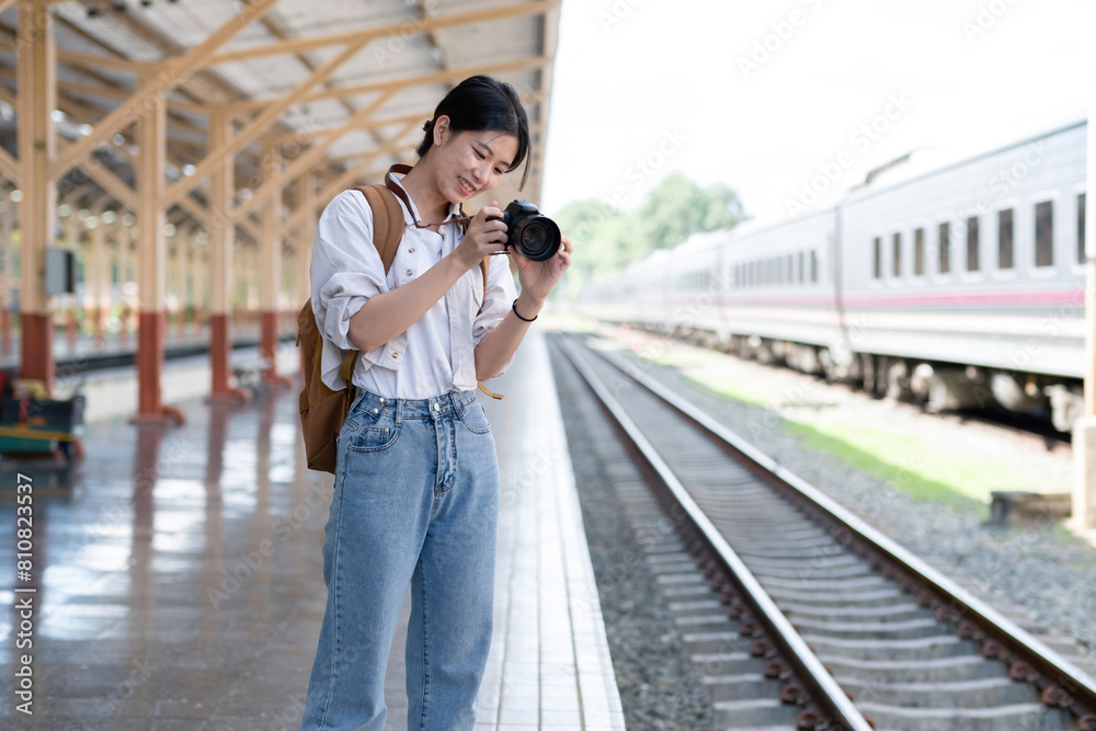 Young Asian woman holds a camera and takes photos of a train while waiting for the train at the train station to travel.