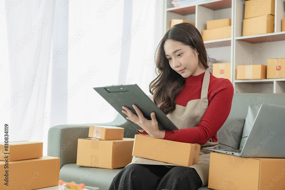 Young Asian business woman smiles and uses laptop to check information on shipping box before sending it to customer. Small business entrepreneur, SME online marketing, delivery concept Young Asian business woman smiles and uses laptop to check information on shipping box before sending it to customer. Small business entrepreneur, SME online marketing, delivery concept