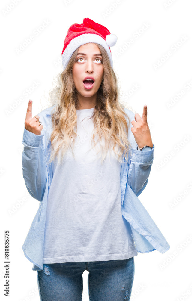 Beautiful young blonde woman wearing christmas hat over isolated background amazed and surprised looking up and pointing with fingers and raised arms. Beautiful young blonde woman wearing christmas hat over isolated background amazed and surprised looking up and pointing with fingers and raised arms.