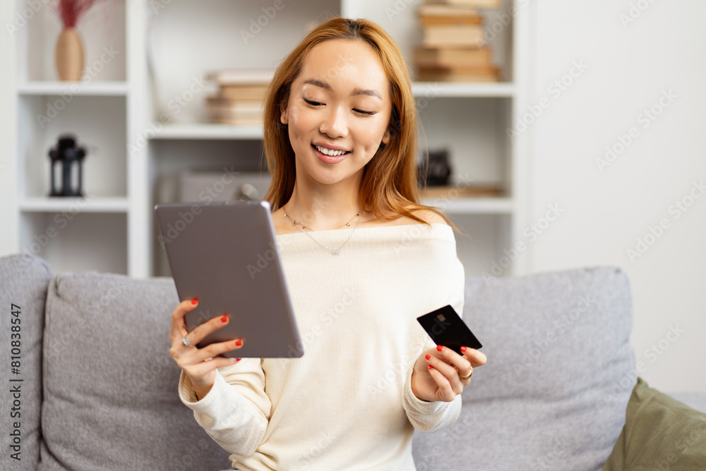 Young Asian Woman Shopping Online On Tablet At Home, Smiling As She Holds A Credit Card, Cozy And Comfortable Living Room Setting Young Asian Woman Shopping Online On Tablet At Home, Smiling As She Holds A Credit Card, Cozy And Comfortable Living Room Setting