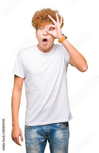 Young handsome man with afro hair wearing casual white t-shirt doing ok gesture shocked with surprised face, eye looking through fingers. Unbelieving expression.