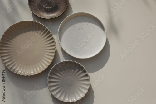 Different size and shapes of plates on beige table background in sunlight. Shadows. Neutral still life, web banner. Food, restaurant, craft concept. Scalloped pottery, ceramics. Flat lay, top view. © tabitazn