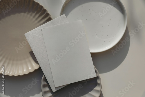 Blank greeting card, invitation mockup, envelope in sunlight. Different plates on beige table background. Soft shadows. Neutral still life, web banner. Food, restaurant, craft concept. Flat lay, top. © tabitazn