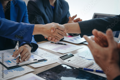 In a business team meeting, success is celebrated with a handshake. Hands close up, dressed in formal suits, working at desks with financial papers, calculators, and laptops.