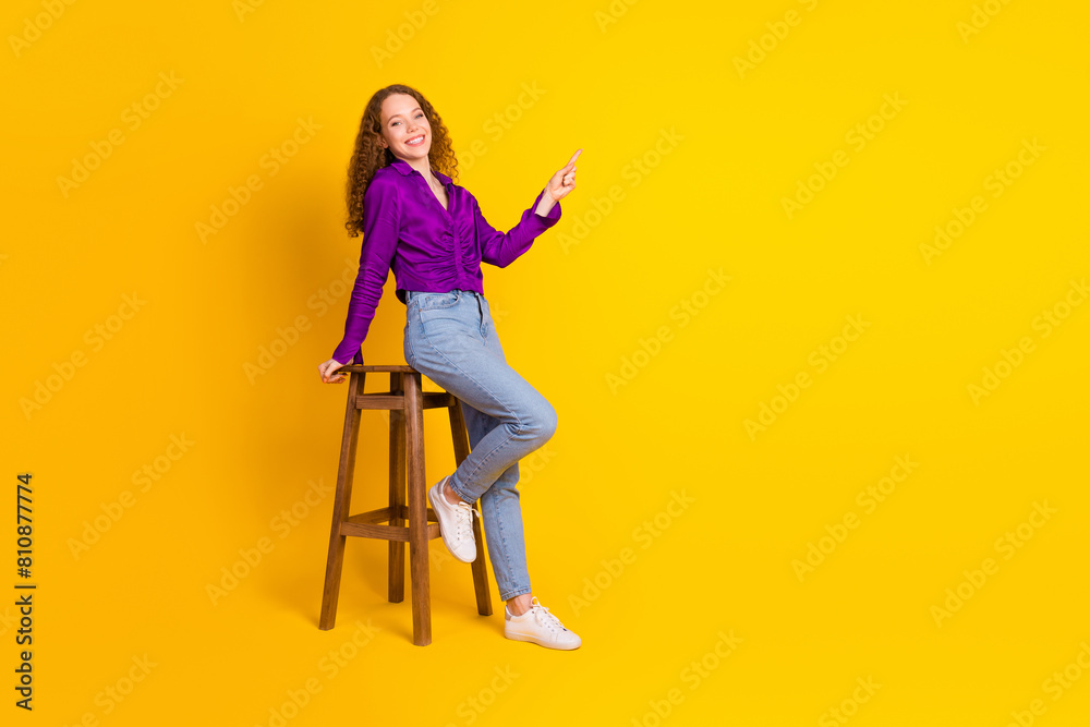 Full body photo of pretty young girl sit chair point empty space wear trendy violet outfit isolated on yellow color background