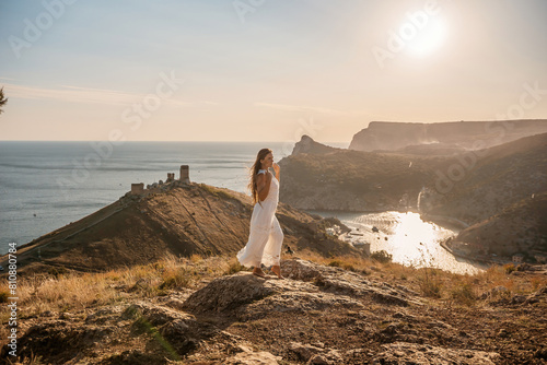 woman stands on a rocky hill overlooking a body of water. She is wearing a white dress and she is in a state of joy or celebration. Concept of freedom and happiness.