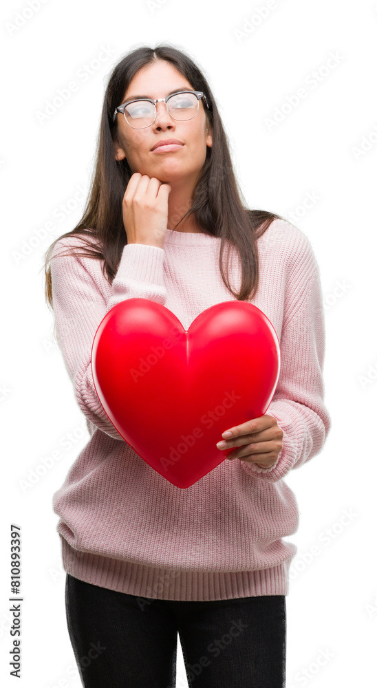Young hispanic woman holding heart in love serious face thinking about question, very confused idea Young hispanic woman holding heart in love serious face thinking about question, very confused idea