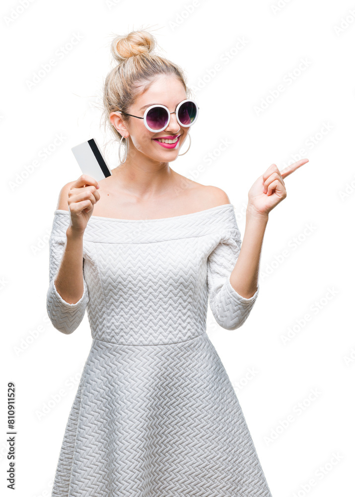 Young beautiful blonde woman holding credit card over isolated background very happy pointing with hand and finger to the side Young beautiful blonde woman holding credit card over isolated background very happy pointing with hand and finger to the side