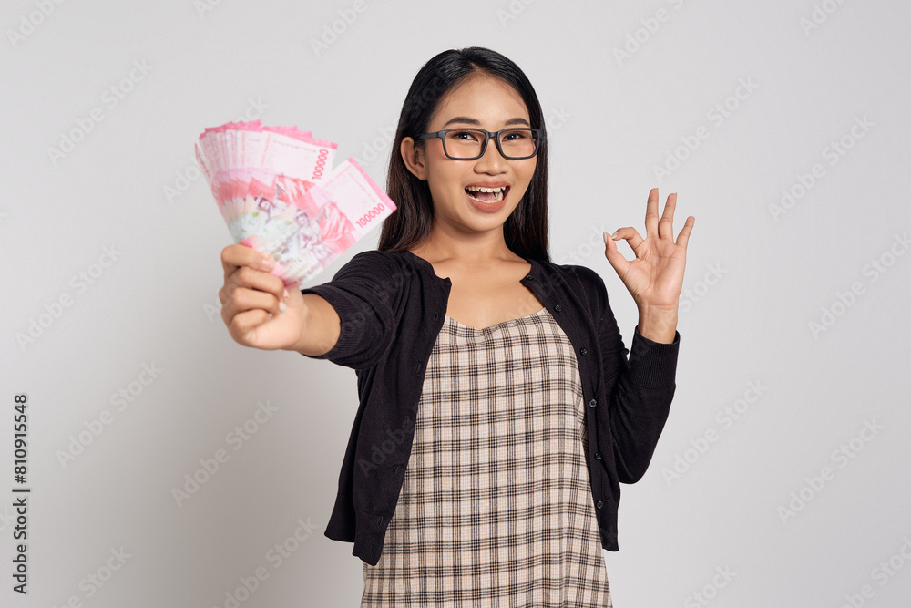 Smiling young Asian woman wearing casual shirt glasses holding money in Indonesian rupiah and showing okay gesture isolated on white background. People lifestyle concept Smiling young Asian woman wearing casual shirt glasses holding money in Indonesian rupiah and showing okay gesture isolated on white background. People lifestyle concept