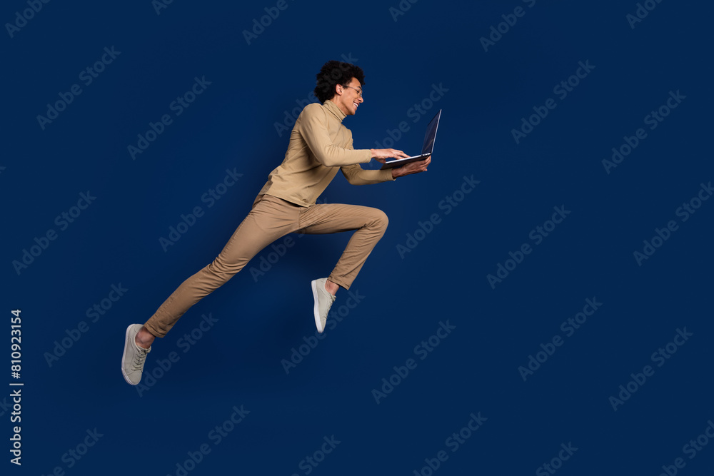 Full size photo of nice young man running fast hold netbook wear trendy beige outfit isolated on dark blue color background Full size photo of nice young man running fast hold netbook wear trendy beige outfit isolated on dark blue color background