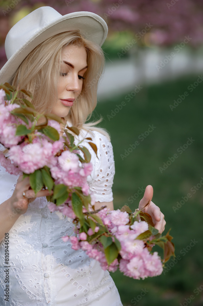 Obraz premium Beautiful young blonde woman in a hat holding a sakura branch in front of her