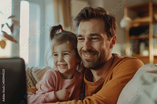 A happy Caucasian father in his 30s sits with his daughter at home, watching an interesting video on a kids' channel online. They cherish the moment together, finding joy in each other's company. The