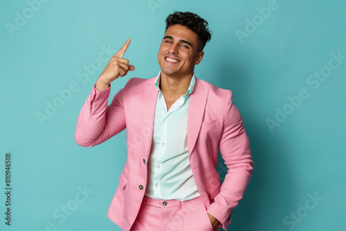 Happy Latino man pointing sideways. Handsome young Latino man in a pink suit standing with his hand in his pocket, isolated on a blue background, showcasing something, advertising a fashion sale,