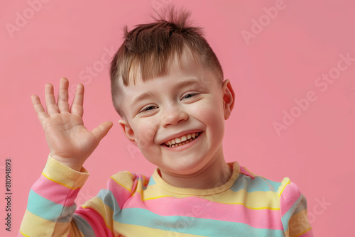 smiling little boy with down syndrome, waving with his hand, wearing striped sweater, on pink background