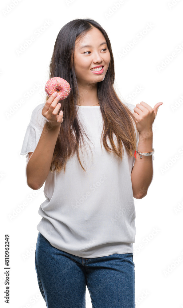 Young asian woman eating donut over isolated background pointing and showing with thumb up to the side with happy face smiling Young asian woman eating donut over isolated background pointing and showing with thumb up to the side with happy face smiling