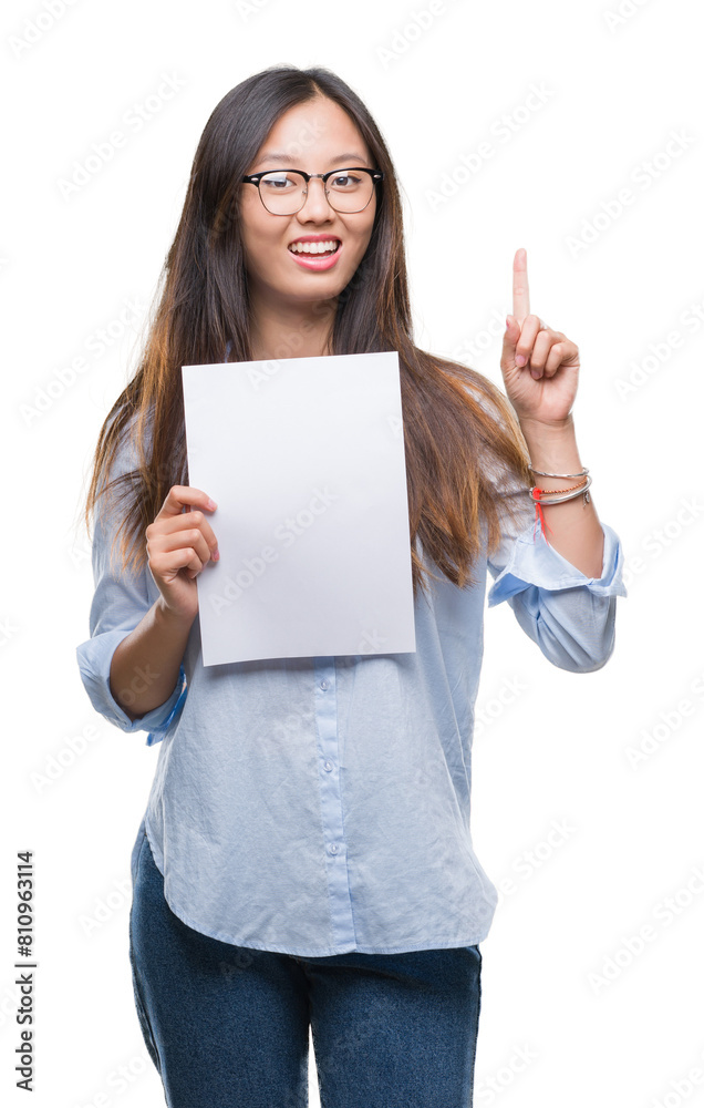Young asian woman holding blank paper over isolated background surprised with an idea or question pointing finger with happy face, number one