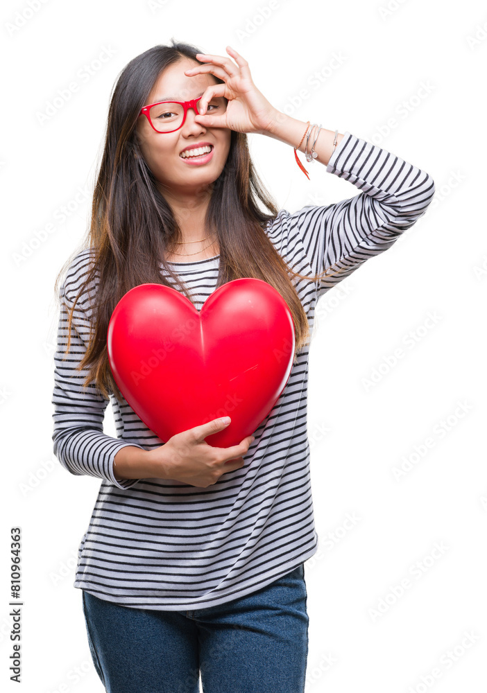Young asian woman in love holding read heart over isolated background with happy face smiling doing ok sign with hand on eye looking through fingers Young asian woman in love holding read heart over isolated background with happy face smiling doing ok sign with hand on eye looking through fingers