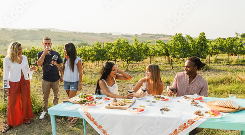 Happy friends having fun drinking red wine and eating together with vineyard in background - Multiracial people doing party at summer time in countryside resort - Main focus on right faces