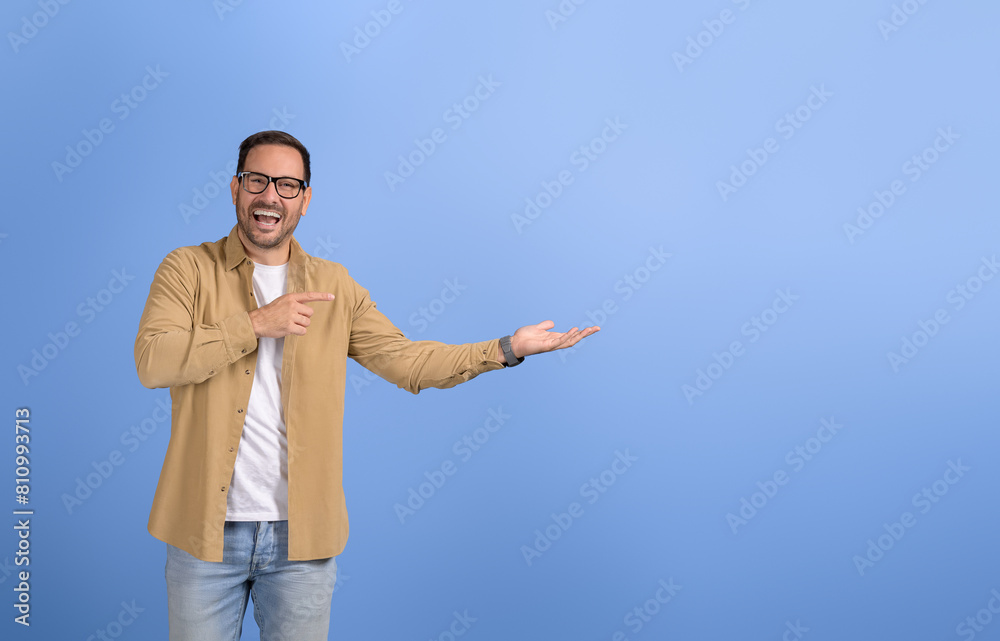 Young businessman laughing and promoting new product on empty palm while standing on blue background Young businessman laughing and promoting new product on empty palm while standing on blue background