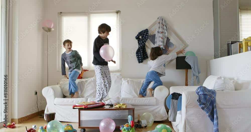 Three kids are energetically jumping on a white couch surrounded by toys and household items in a disordered living room, with sunlight streaming through the window.