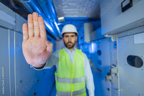 Electrical engineer technician in protective vest and white helmet stretch out his palm signaling danger. Substation worker say stop backdrop with blue light high voltage, faulty electrical equipment.