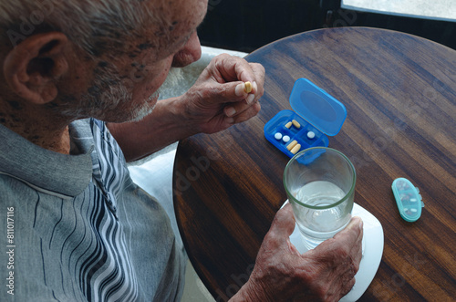 Hombre mayor nonagenario sentado sosteniendo una píldora y un vaso de agua con pastillero sobre mesa de madera. Plano picado.  photo