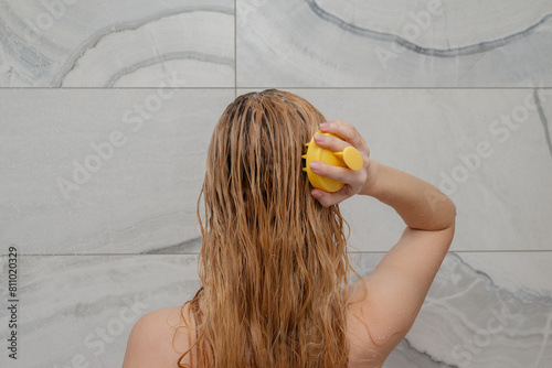 Blonde girl with long hair with her back turned washes her hair in the shower and does a self-massage of her head with a massage brush. Self-care, home spa, scalp cleansing.