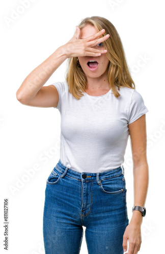 Beautiful young woman wearing casual white t-shirt over isolated background peeking in shock covering face and eyes with hand, looking through fingers with embarrassed expression. © Krakenimages.com