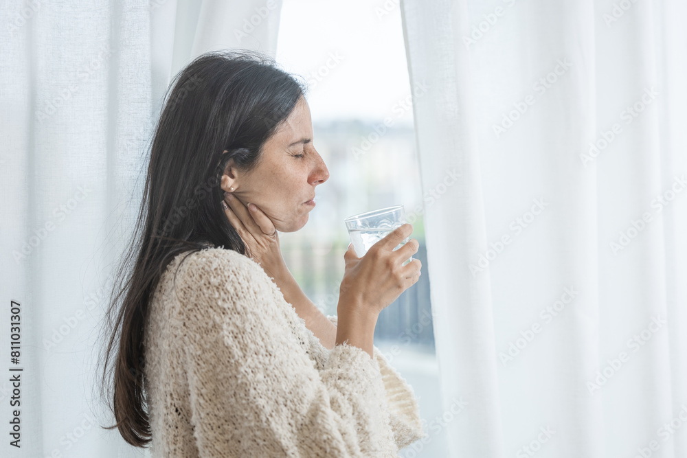 Woman near window holding glass of medicine for sore throat. Overwhelmed woman suffering from sore throat. Woman near window holding glass of medicine for sore throat. Overwhelmed woman suffering from sore throat.