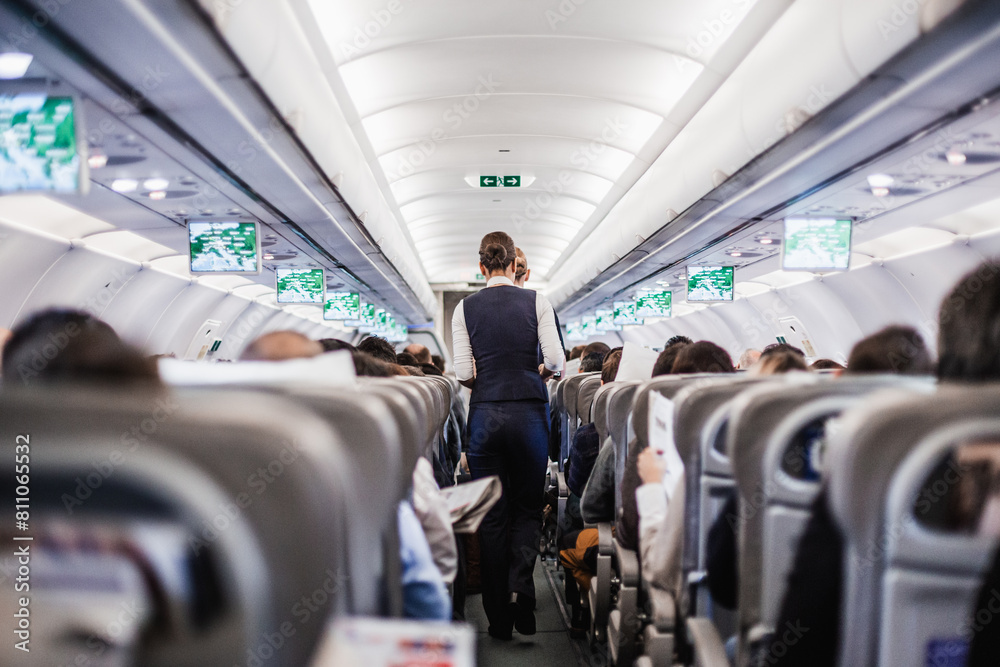 Interior of airplane with passengers on seats and stewardess in uniform walking the aisle, serving people. Commercial economy flight service concept Interior of airplane with passengers on seats and stewardess in uniform walking the aisle, serving people. Commercial economy flight service concept