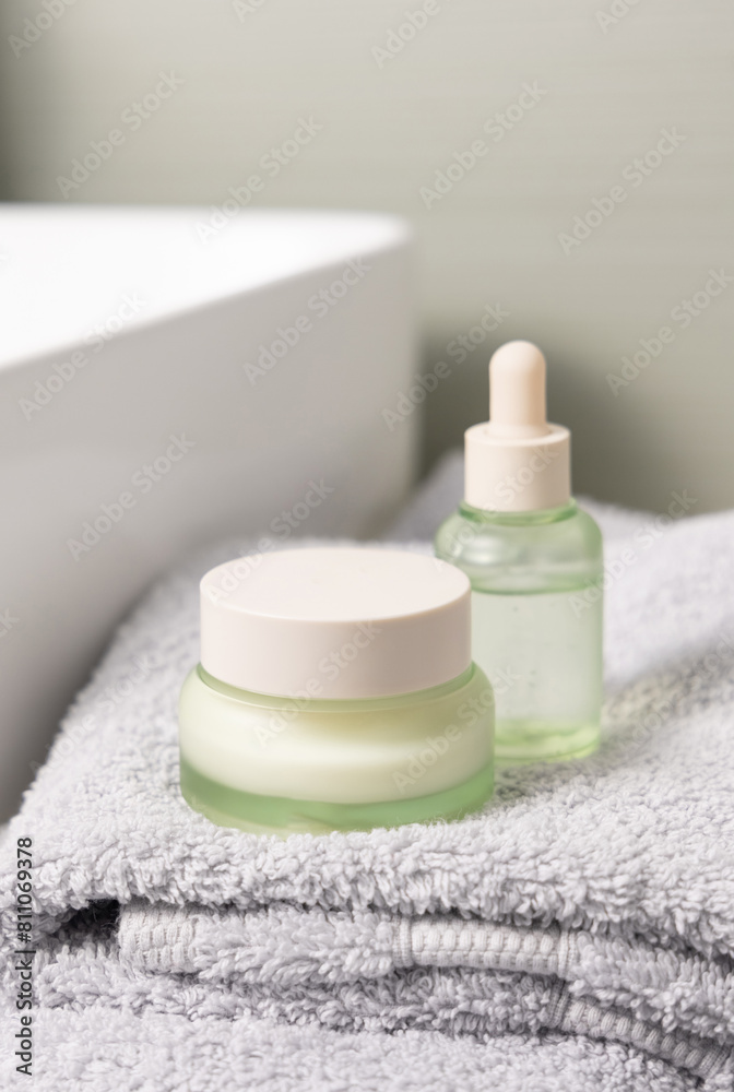 Light green dropper bottle and cream jar on light grey bath towel near basin closeup Light green dropper bottle and cream jar on light grey bath towel near basin closeup
