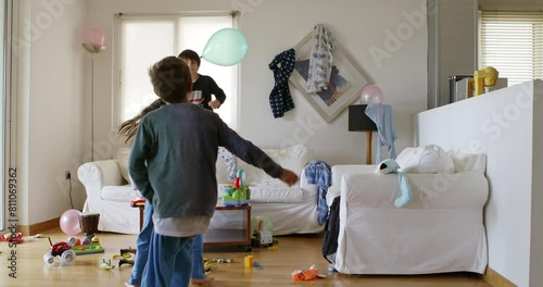 Three kids are energetically jumping on a white couch surrounded by toys and household items in a disordered living room, with sunlight streaming through the window.