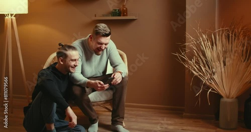 Cute Male Gay Couple Spend Time. Sitting chair floor Use Smartphone. Browse Online laughing. Partner's Hand is Around His Lover. They Smile and Laugh. Room Modern Interior. shopping together from home photo