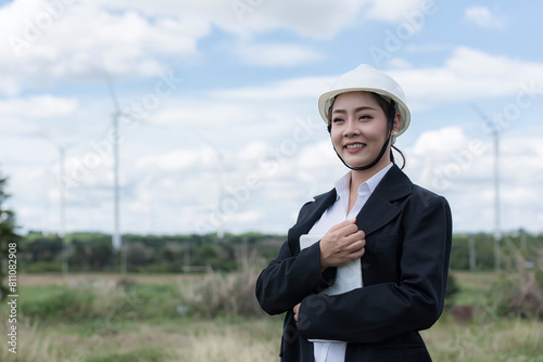 businesswoman with arms crossed. Beautiful Asian woman in white helmet working with digital tablet at renewable energy farm. Female inspector controlling functioning of wind turbines outdoors.