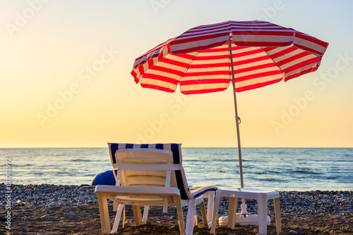 A beach scene with a red and white striped umbrella and a white beach chair