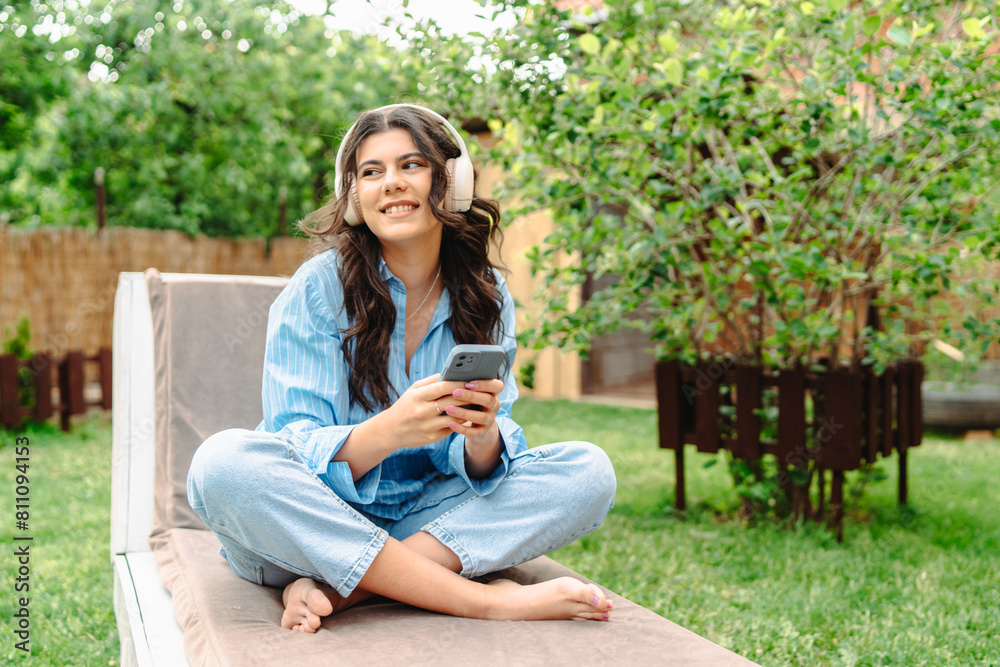 One young caucasian woman is relaxing in easy chair in her backyard while using her phone and listening to music on wireless headphones One young caucasian woman is relaxing in easy chair in her backyard while using her phone and listening to music on wireless headphones