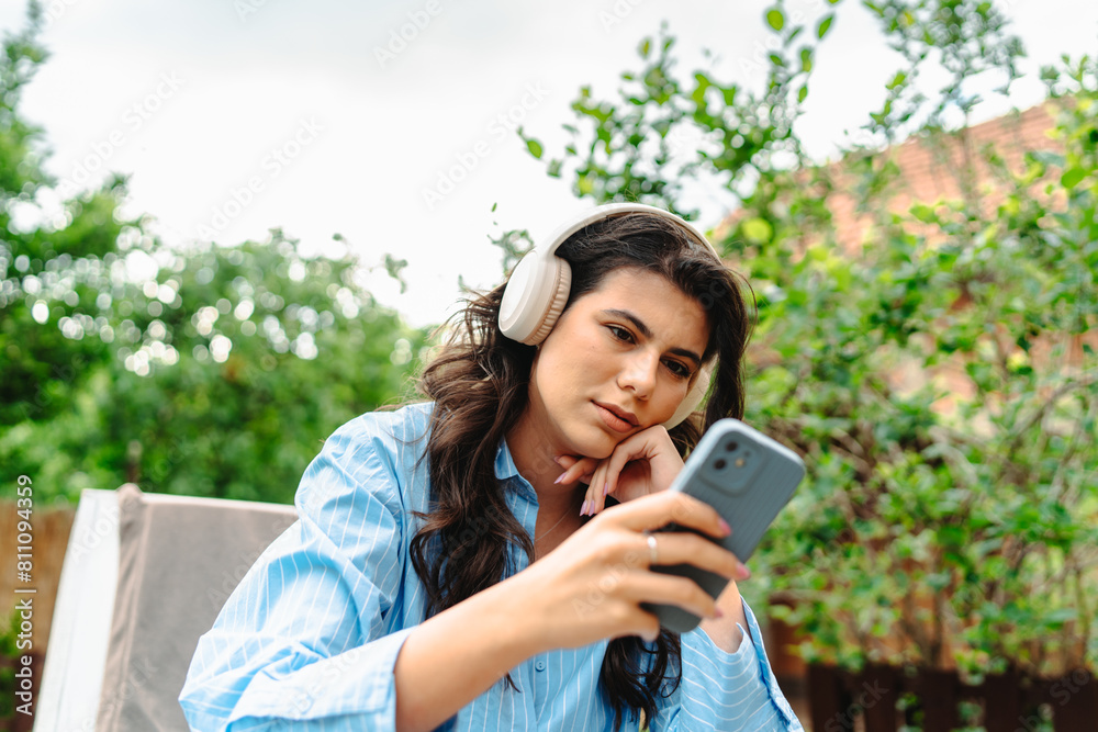 One young caucasian woman is relaxing in easy chair in her backyard while using her phone and listening to music on wireless headphones