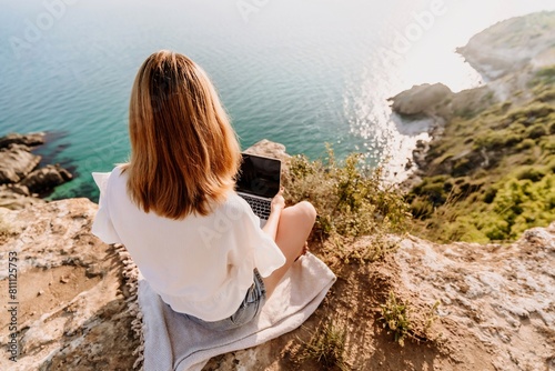 A woman is sitting on a rock overlooking the ocean with a laptop in front of her. She is enjoying the view and the peacefulness of the location.