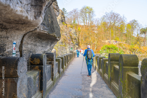 A senior traveler with a backpack and trekking poles walks on the Bastei Bridge amid sandstone formations in Saxon Switzerland National Park. Germany