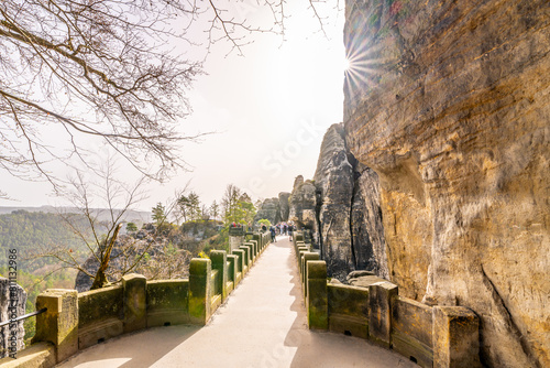 Visitors walk along the historic Bastei Bridge, bathed in sunlight, amidst the rock formations of Saxon Switzerland National Park. Germany