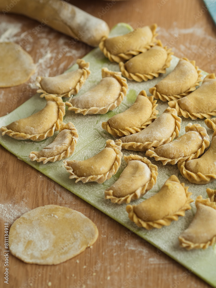 Preparing dumplings: A vertical close-up of Ukrainian varenyky on a cutting board, arranged in three rows. Dough circles are visible, with flour scattered across the table. Preparing dumplings: A vertical close-up of Ukrainian varenyky on a cutting board, arranged in three rows. Dough circles are visible, with flour scattered across the table.