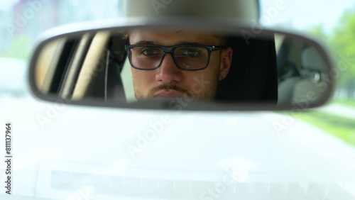 Smiling young man reflected in car rear view mirror. Happy guy in glasses driving car in the city. Transport, business, lifestyle and people concept. Real time