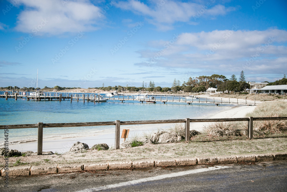 A road runs alongside a Thomson Bay, Rottnest Island, offering a view of the serene bay and jetty. The water reflects the sky above, creating a peaceful scene. A road runs alongside a Thomson Bay, Rottnest Island, offering a view of the serene bay and jetty. The water reflects the sky above, creating a peaceful scene.