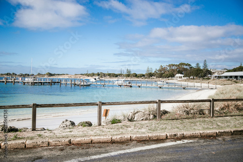 A road runs alongside a Thomson Bay, Rottnest Island, offering a view of the serene bay and jetty. The water reflects the sky above, creating a peaceful scene.